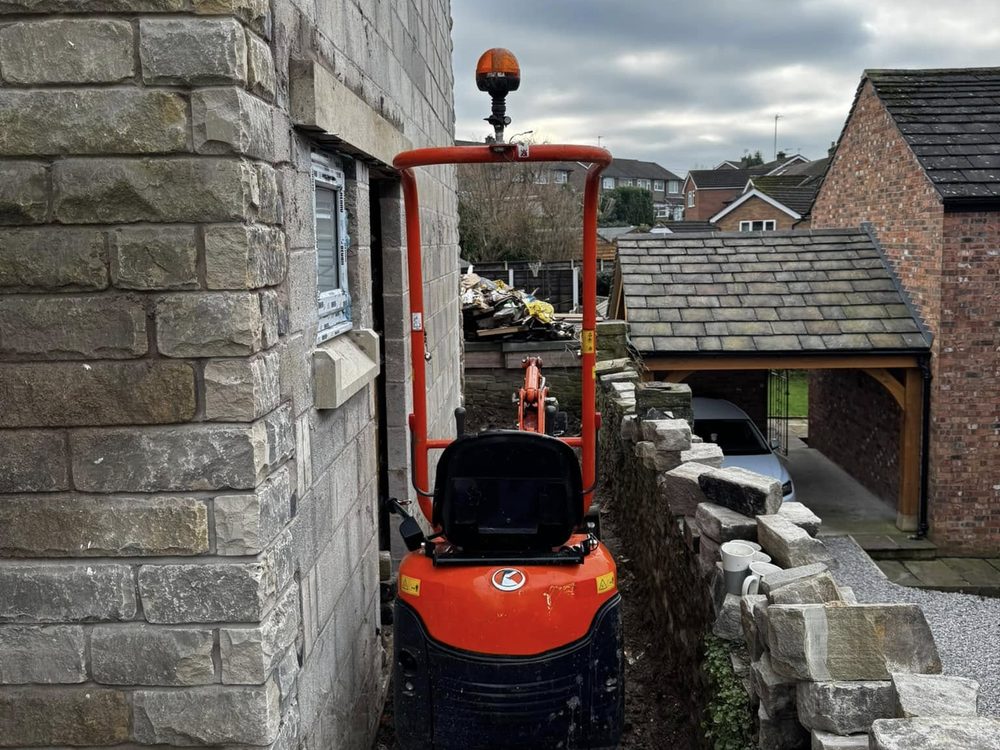 Kubota micro digger working down a narrow side passage between a house and a stone wall